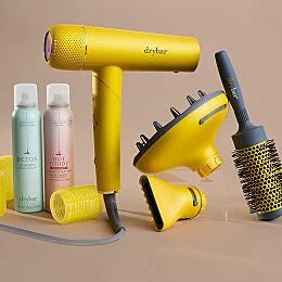 Drybar Buttercup Pro hair dryer displayed with styling attachments, round brush, hair rollers, dry shampoo, and heat protectant spray.